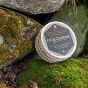 A tin of Killiecranking Recovery Cream, its lid adorned with descriptive text, rests gracefully on moss-covered rocks. Trail Science Recovery Cream
